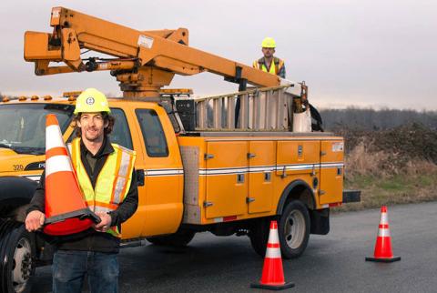 Man with a hard hat and high-visibility vest holds an orange traffic cone, with a work truck in the background.